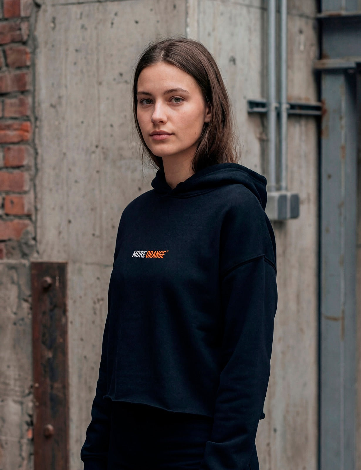 Side view of female model wearing black MORE ORANGE™ hoodie in minimalist urban style