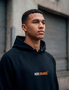 Male model wearing black MORE ORANGE™ hoodie photographed in urban concrete setting