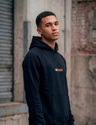 Male model wearing black MORE ORANGE™ hoodie standing in minimalist city environment